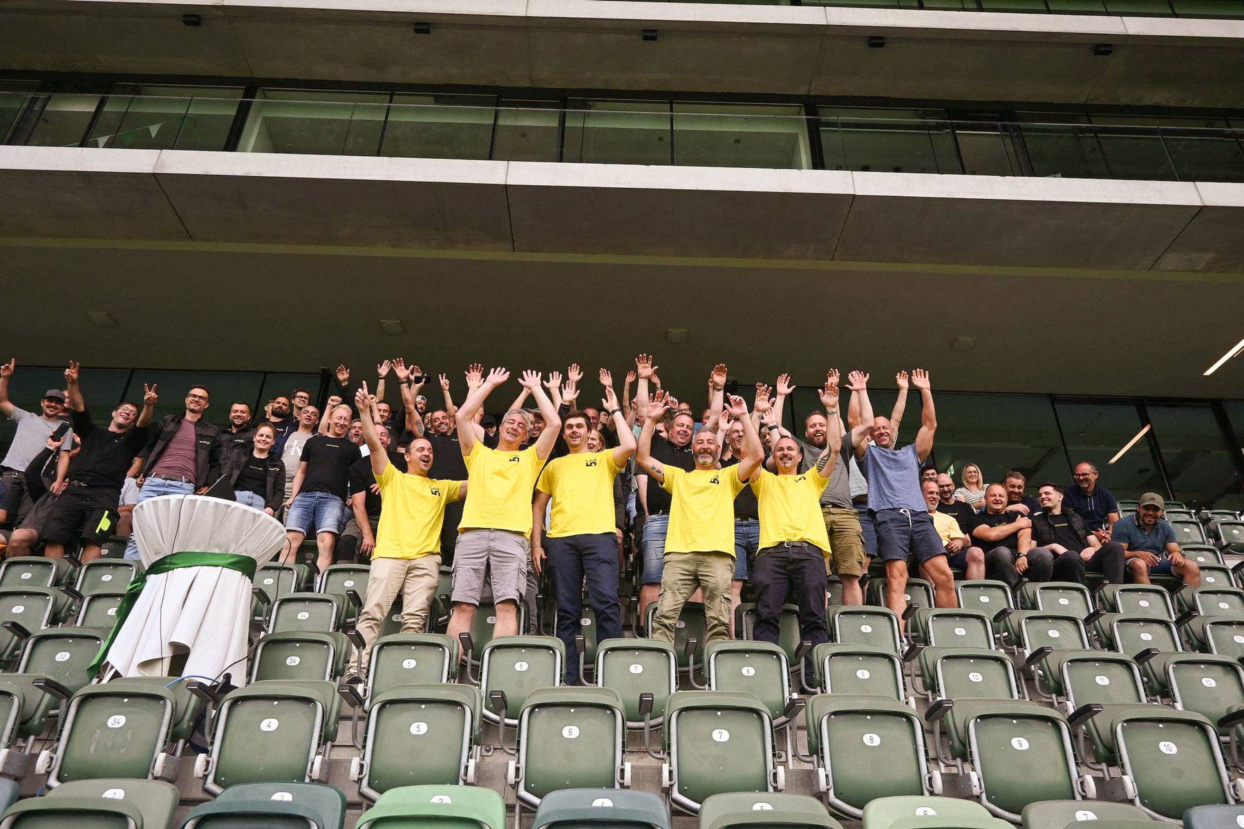 5er Clan: Team in gelben Shirts auf der Stadiontribüne